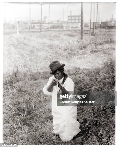 View of an unidentified young woman, in a hat and a light-colored dress, as she kneels on one leg and aims a BB gun, Lincoln, Nebraska, early 20th...