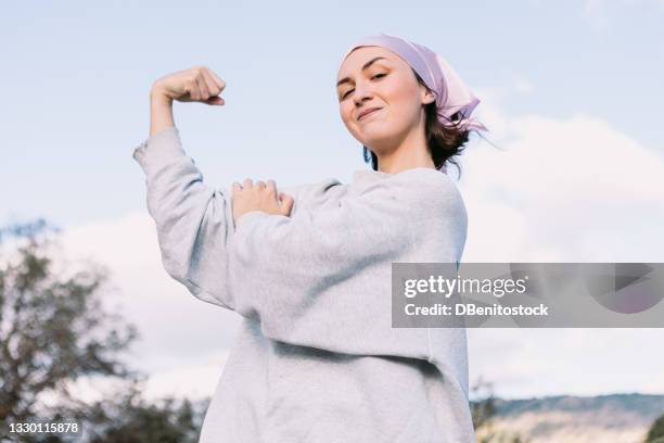 young girl sick with cancer, in the field, making the sign of strength with her arms, wearing a pink scarf on her head, international breast cancer day, with the sky in the background. breast cancer concept - cancer survivor stockfoto's en -beelden