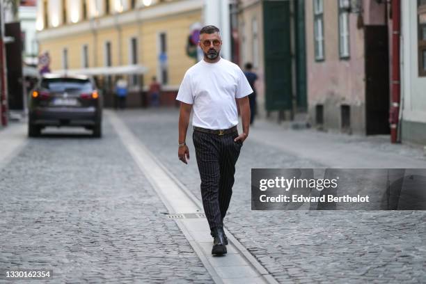 Guest wears sunglasses, a white t-shirt, a brown leather belt with golden buckle detail, gray and white striped suit pants, leather boots, during...