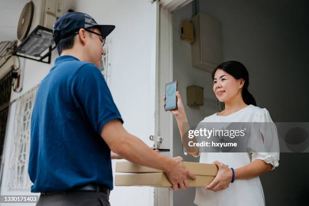 young asian woman receiving a parcel and making payment with mobile phone - pizza delivery stock pictures, royalty-free photos & images