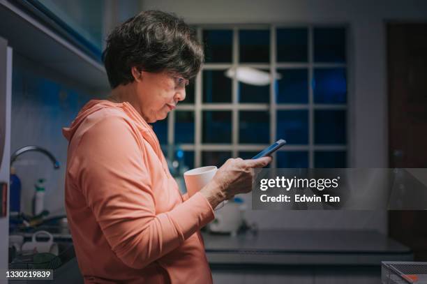 side view asian chinese senior woman leaning and using smart phone and drinking coffee in domestic kitchen at night - phone message stock pictures, royalty-free photos & images