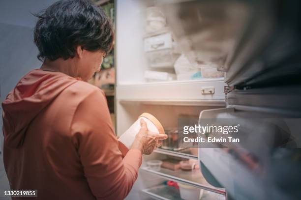 vista trasera asiática china senior mujer abriendo refrigerador sacando helado congelado por la noche en la cocina - congelador fotografías e imágenes de stock