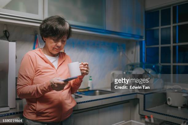 asian chinese senior woman leaning and using smart phone and drinking coffee in domestic kitchen at night - old woman by window stock pictures, royalty-free photos & images