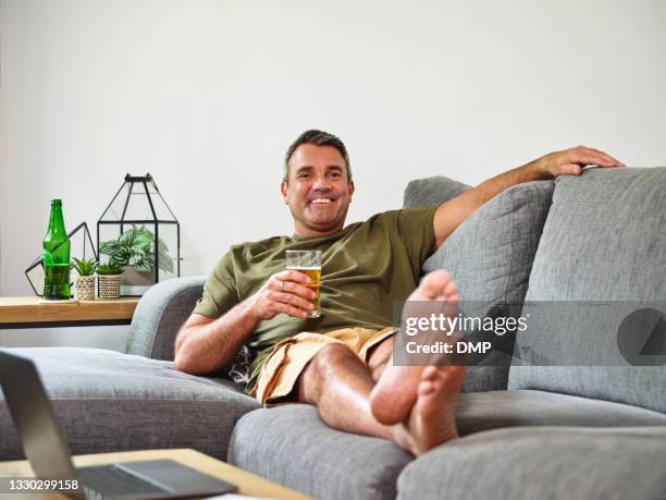 shot of a mature man having a glass of beer on the sofa at home - man sipping beer smiling stockfoto's en -beelden