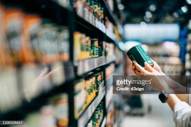 close up of a woman grocery shopping in supermarket. holding a tin can and reading the nutrition label at the back - consumerism foto e immagini stock