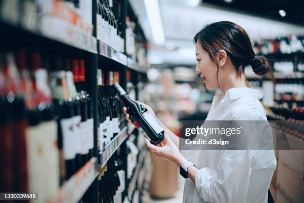 young asian woman walking through liquor aisle and choosing a bottle of red wine from the shelf in supermarket - bottle shop stock pictures, royalty-free photos & images