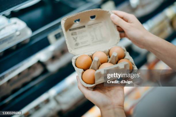 close up of young asian woman grocery shopping in a supermarket. she is holding a box of fresh organic free range eggs in front of a refrigerated section. healthy eating lifestyle - carton of eggs stockfoto's en -beelden