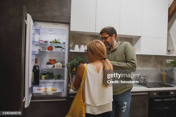 husband and wife putting groceries in the fridge together - opbergen stockfoto's en -beelden