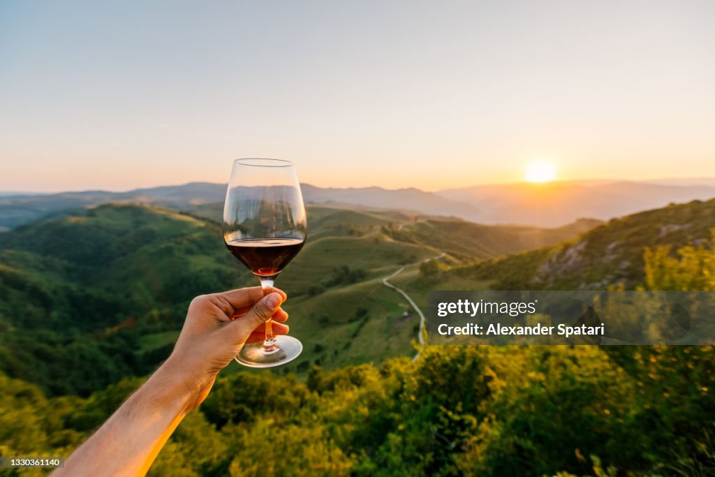Man holding a glass of red wine surrounded by hills and mountains at sunset, personal perspective POV