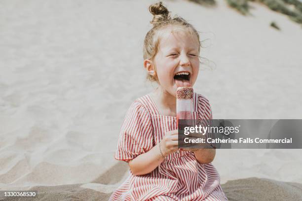 cute girl licks a ice lolly, while sitting on the beach - freezing hands stockfoto's en -beelden