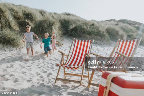 children joyfully run down a sand dune towards deckchairs and a cool box - vilfåtölj bildbanksfoton och bilder