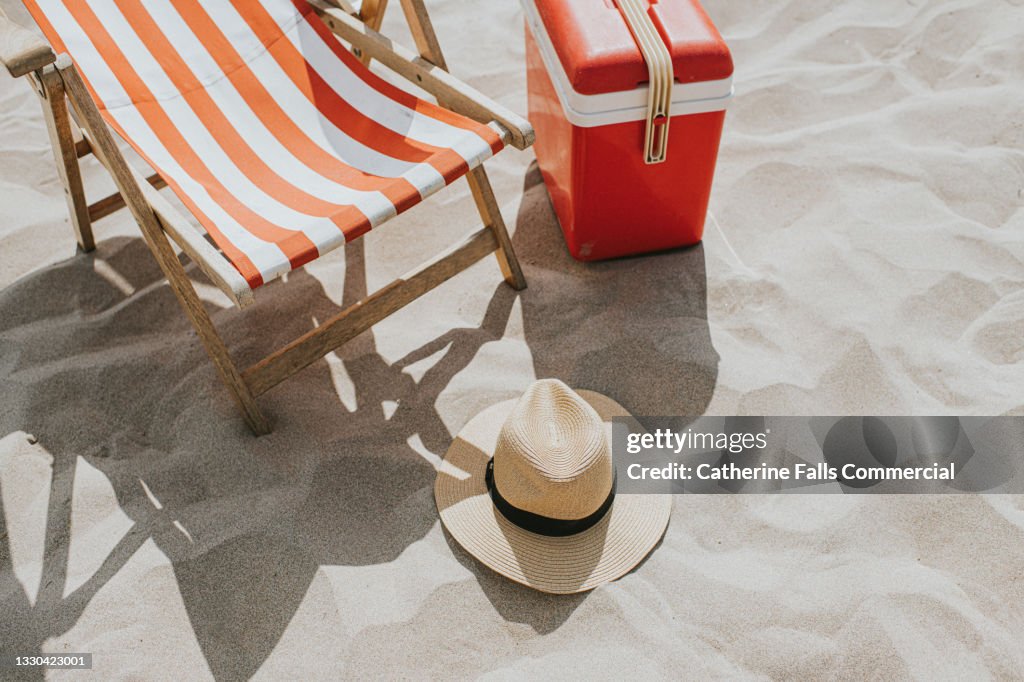 Overhead view of a relaxing beach scene, including a deck chair, sun hat and cool box.