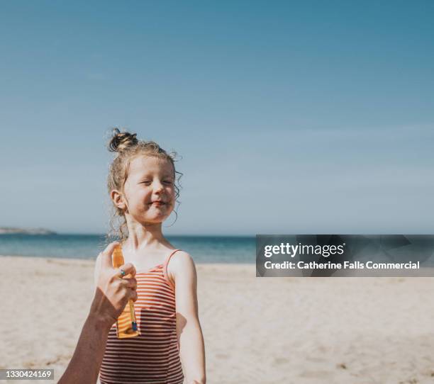 a cute little girl half smiles and grimaces as an adult applies sun block to her face - protection humour stock-fotos und bilder