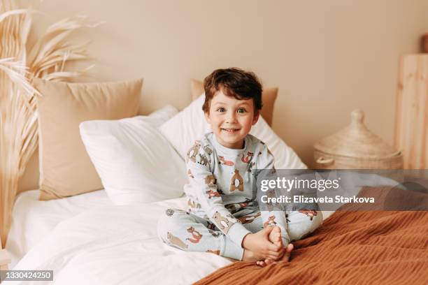 portrait of a happy cheerful little boy a teenage child in night pajamas with dinosaurs sitting on a cozy bed and looking at the camera in a comfortable bedroom at home on a day off on vacation - pyjama stockfoto's en -beelden