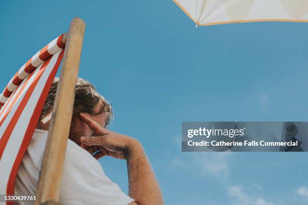 man sits in a striped deckchair against blue sky under parasol - hittegolf stockfoto's en -beelden