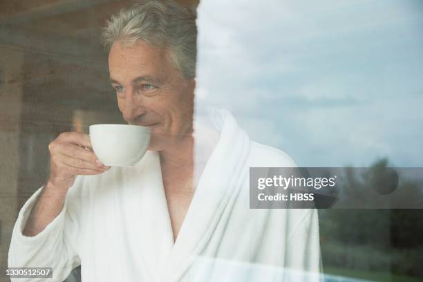 senior man in bathrobe behind a window drinking from cup - bathrobe stock pictures, royalty-free photos & images