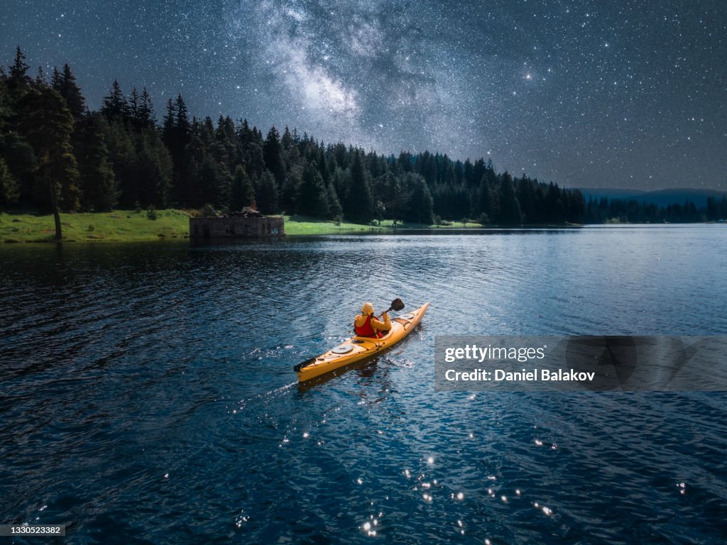 Mujer en kayak en el lago de montaña bajo la Vía Láctea. Vista aérea con cielo estrellado. Remo y ecoturismo.