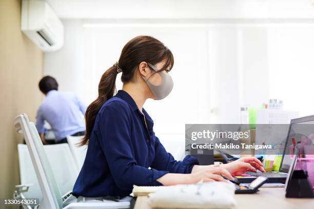 young japanese woman wearing a mask and working at the office during the coronavirus pandemic - businesswoman mask stock pictures, royalty-free photos & images