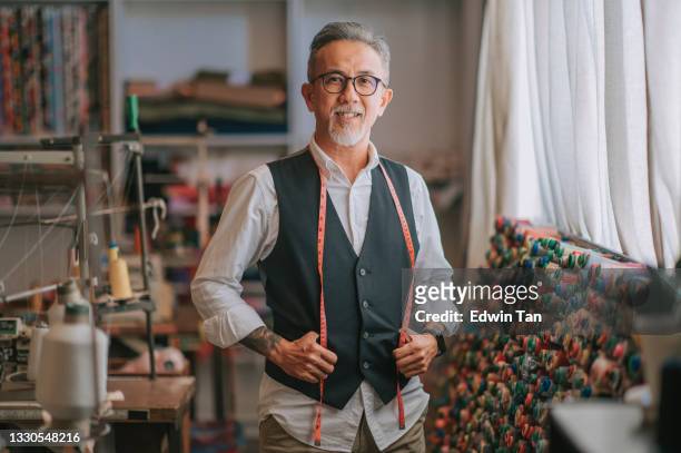 asian chinese senior tailor man with facial hair looking at camera smiling in atelier studio - atelier fashion stock pictures, royalty-free photos & images