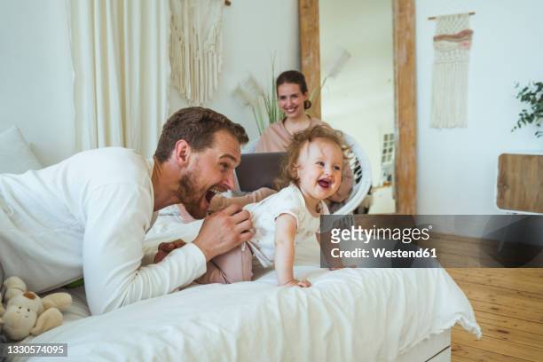 smiling woman looking at cheerful man playing with daughter in bedroom - crawling stock pictures, royalty-free photos & images