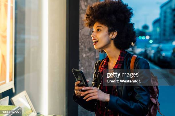 surprised woman holding mobile phone doing window shopping at dusk - montra imagens e fotografias de stock