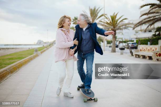 cheerful mature man skateboarding by woman walking while holding hands on footpath - couple skating stock pictures, royalty-free photos & images