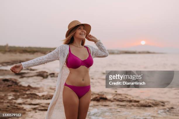 hermosa joven con un bikini con los brazos abiertos y sosteniendo su sombrero de verano de pie con una gran sonrisa mirando hacia la playa durante la puesta de sol - vista marina fotografías e imágenes de stock