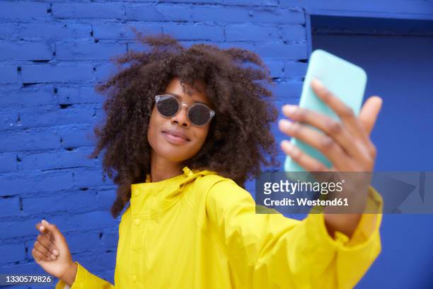 young woman wearing sunglasses taking selfie through smart phone in front of blue wall - holding sunglasses stock pictures, royalty-free photos & images
