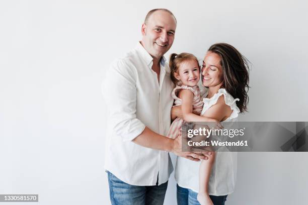 smiling man standing by woman carrying daughter in front of white background - child portrait studio stock-fotos und bilder