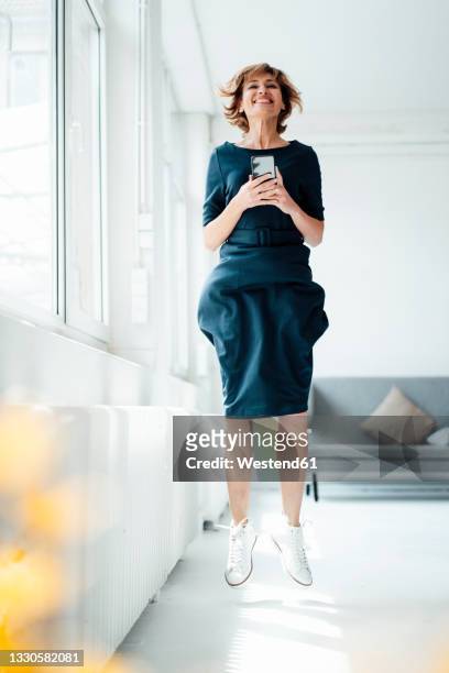 businesswoman holding mobile phone while jumping in office - frau springen stock-fotos und bilder