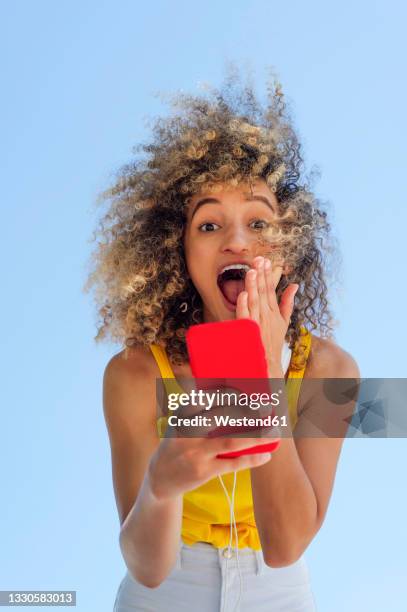 surprised woman with curly hair using mobile phone - stupore esterno foto e immagini stock