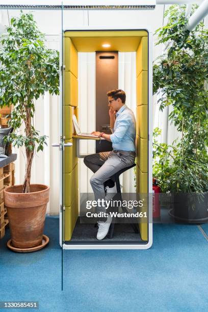 male professional talking on mobile phone while using laptop in soundproof booth - booth foto e immagini stock