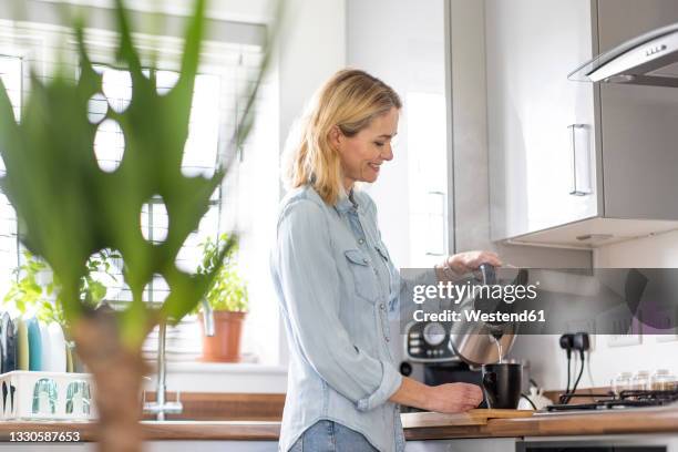 smiling woman pouring water in mug while preparing tea in kitchen at home - やかん ストックフォトと画像