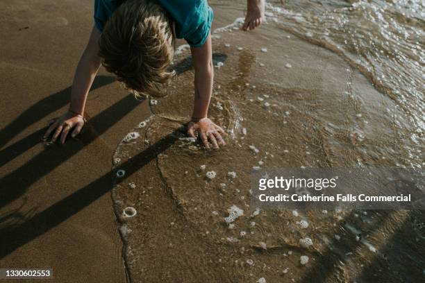 child paddles into the ocean - bending over to dip his hands into the shallow tide - rippled sand stock pictures, royalty-free photos & images