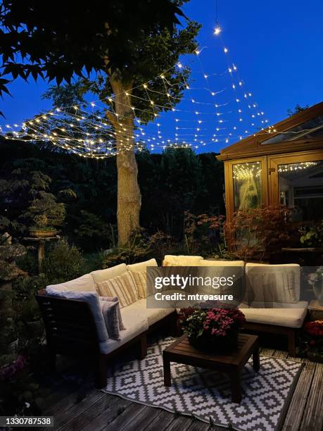 image de la zone de détente extérieure la nuit éclairée par des lumières de fées à cordes, des sièges en bois franc avec coussins, une table en bois avec une pièce maîtresse de plante à fleurs, des bonsaïs, des érables japonais, un jardin de d - garden night photos et images de collection