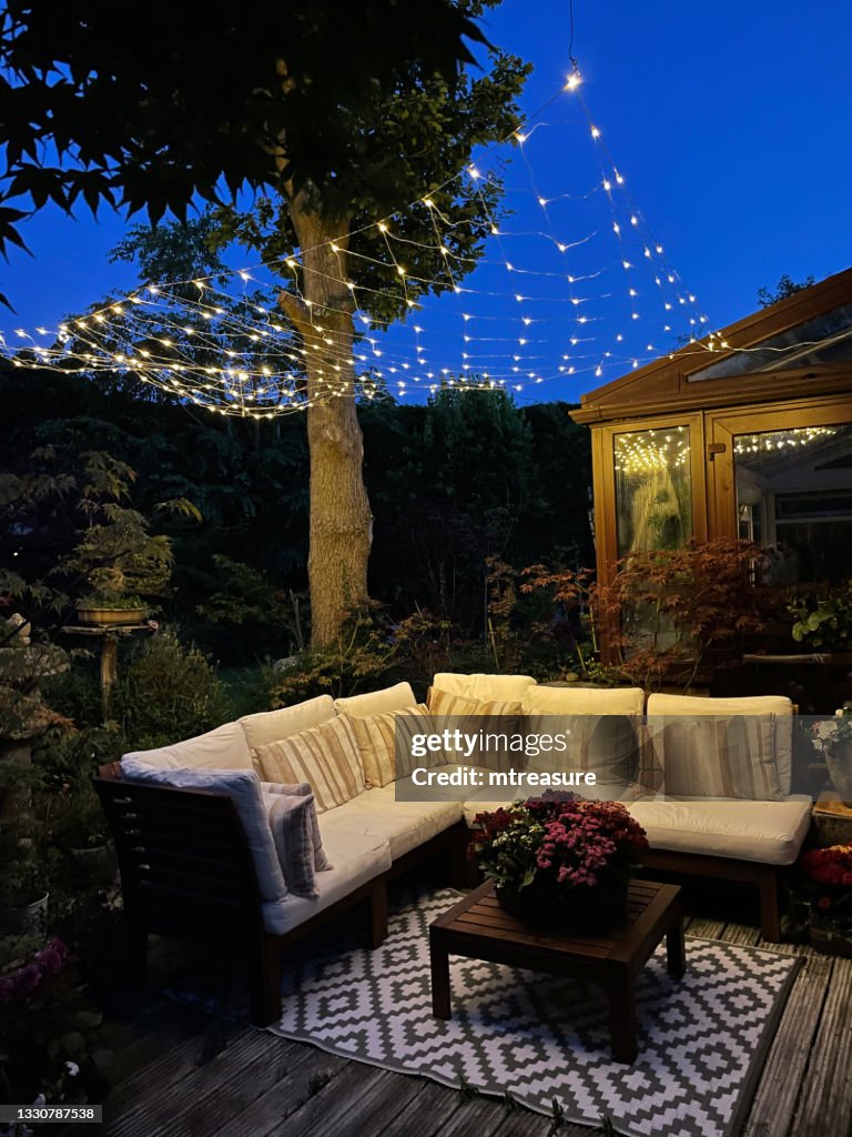 Image of outdoor lounging area at night illuminated by string fairy lights, hardwood seating with cushions, wooden table top with flowering plant centrepiece, bonsai trees, Japanese maples, landscaped oriental design garden, focus on foreground