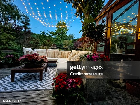 Imagen de la zona de descanso al aire libre por la noche iluminada por luces de hadas de cuerda, asientos de madera dura con cojines, mesa de madera con pieza central de plantas con flores, árboles de bonsái, arces japoneses, jardín de diseño oriental Imagen de la zona de descanso al aire libre por la noche iluminada por luces de hadas de cuerda, asientos de madera dura con cojines, mesa de madera con pieza central de plantas con flores, árboles de bonsái, arces japoneses, jardín de diseño oriental