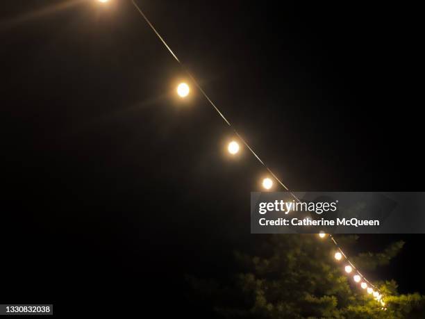 festive hanging string of lights against a dark black summer sky - cadena de luces fotografías e imágenes de stock
