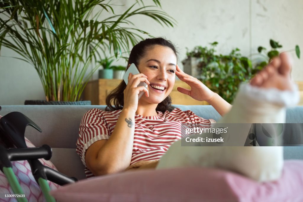Cheerful young woman with broken leg on phone