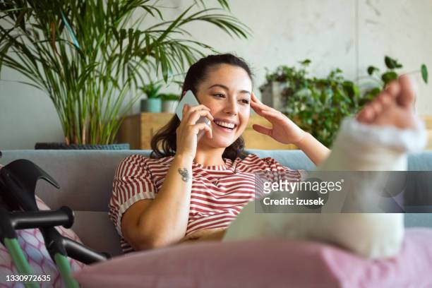 cheerful young woman with broken leg on phone - accident recovery stockfoto's en -beelden