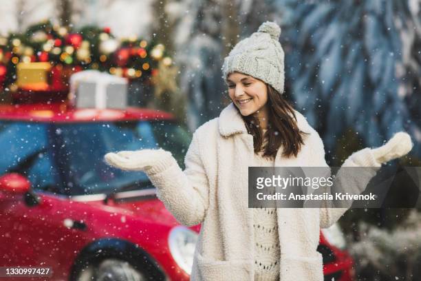 positive woman in wool mittens and fur jacket standing near car with green christmas tree and presents on top. - knitted car stock pictures, royalty-free photos & images