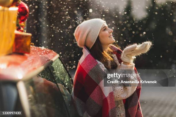 positive woman in wool mittens and fur jacket standing near car with green christmas tree and presents on top. - frau geschenk stock-fotos und bilder