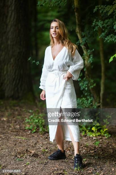 Guest wears a white V-neck long sleeves linen long belted wrap dress, black leather and fabric sneakers, , on July 22, 2021 in Sibiu, Romania.