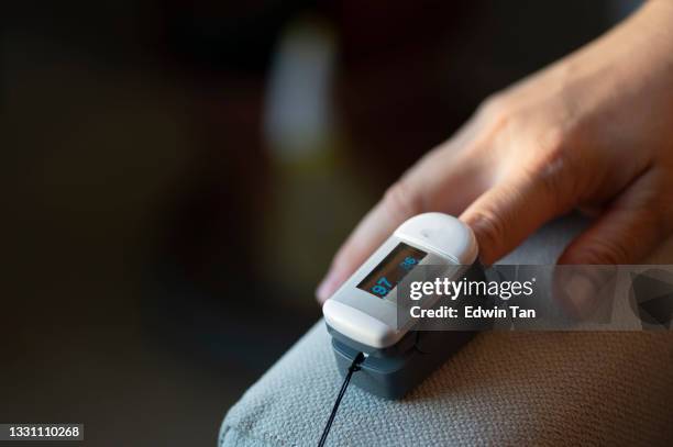 close up asian chinese mid adult woman's finger attached with oximeter measuring oxygen saturation in blood and heartbeat rate at home covid-19 health technology - pulse oximeter stock pictures, royalty-free photos & images