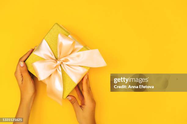 woman hands with manicure are holding gold gift box with silk beige bow on illuminating yellow background. trendy colors of the year 2021. christmas celebration concept. flat lay style. copy space for your design - surprise gift foto e immagini stock
