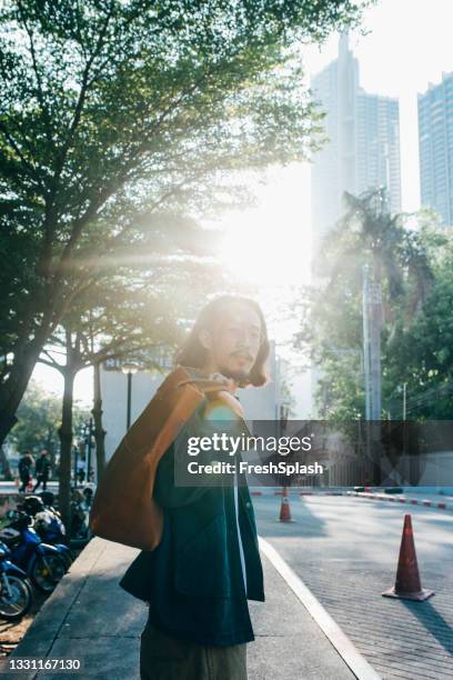 handsome thai man carrying a bag over his shoulder and crossing the street - over shoulder man stockfoto's en -beelden