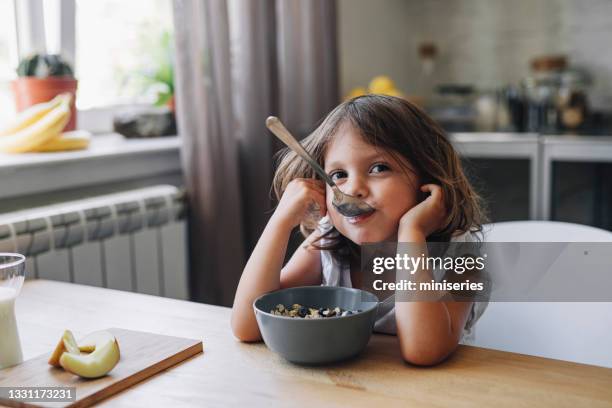 süßes kleines mädchen, das in die kamera schaut, während es müsli zum frühstück zu hause isst - children eating breakfast stock-fotos und bilder