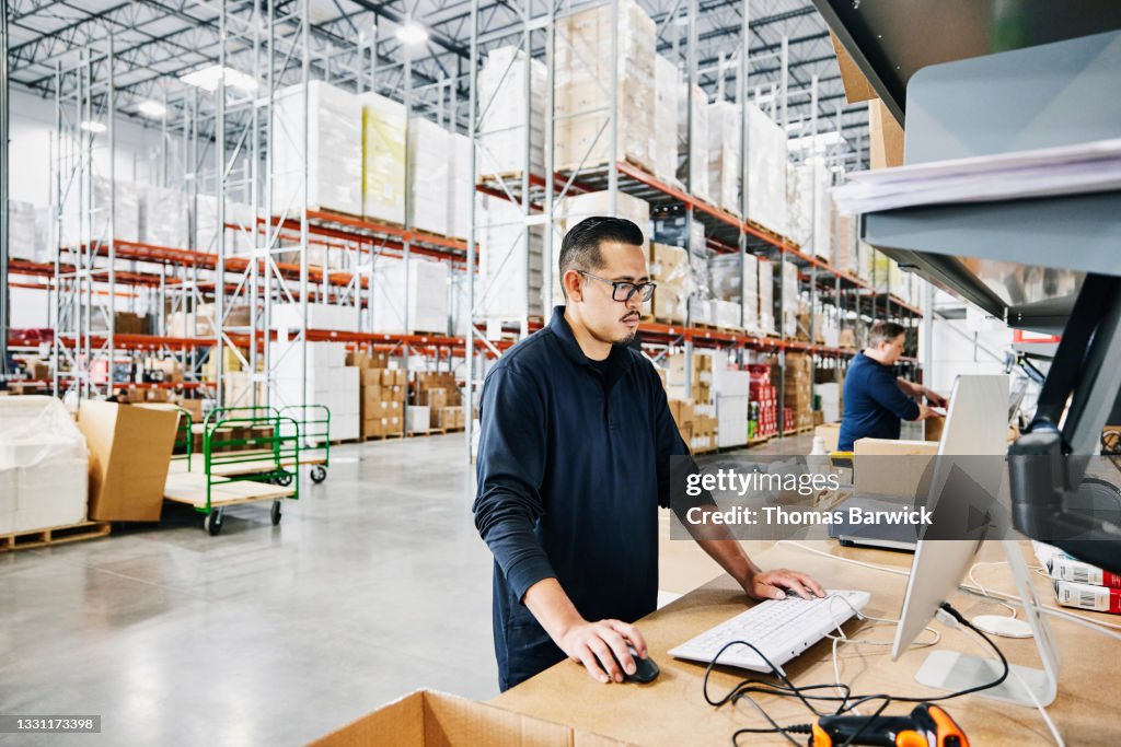 Medium wide shot of male warehouse worker checking orders at computer workstation in warehouse