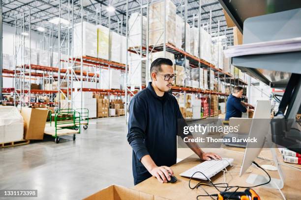 medium wide shot of male warehouse worker checking orders at computer workstation in warehouse - years of export stockfoto's en -beelden