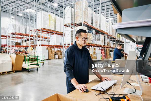Medium wide shot of male warehouse worker checking orders at computer workstation in warehouse
